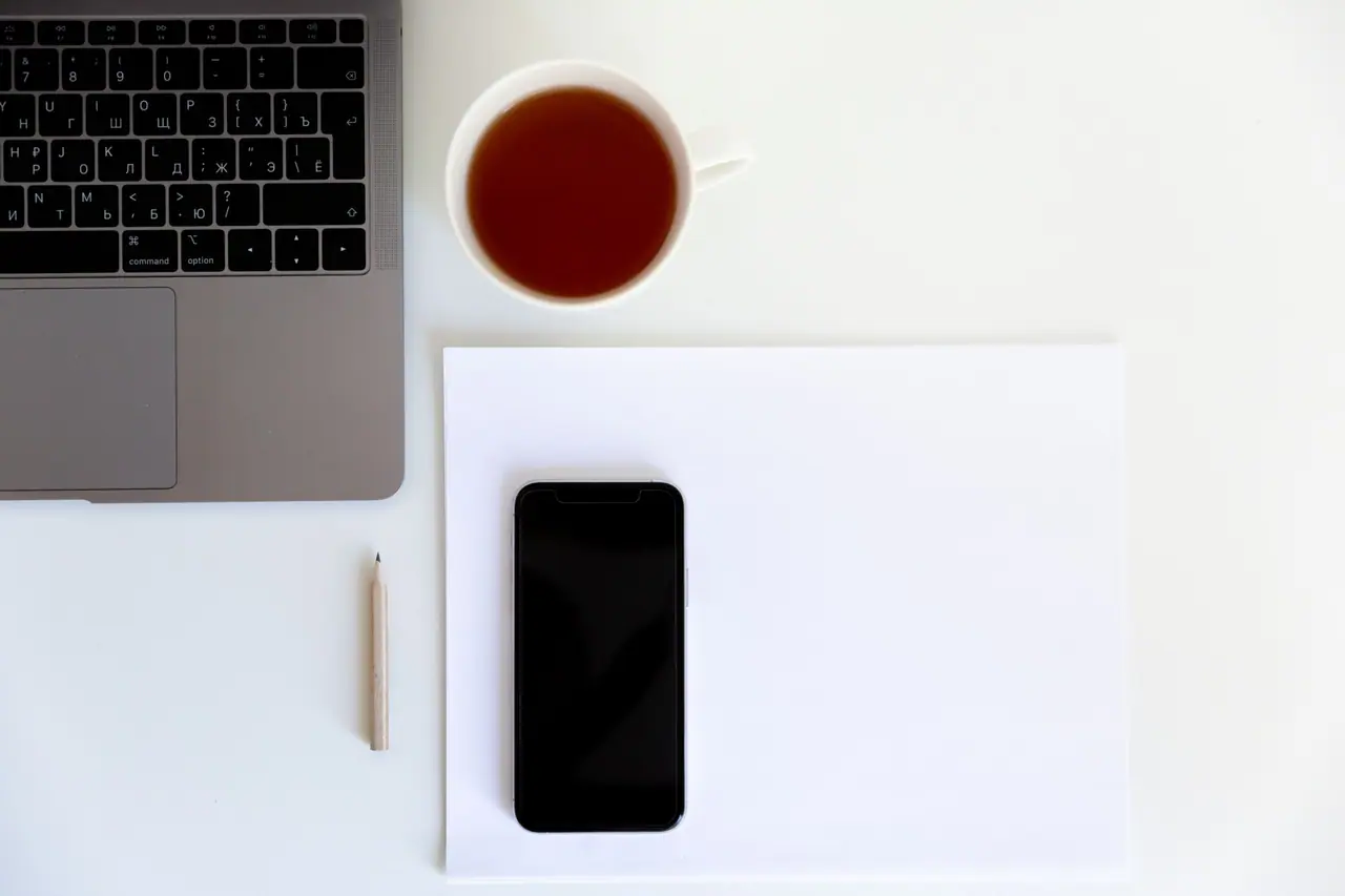 Sur un bureau vu de dessus, un téléphone posé sur une feuille, une tasse et un ordinateur portable.
